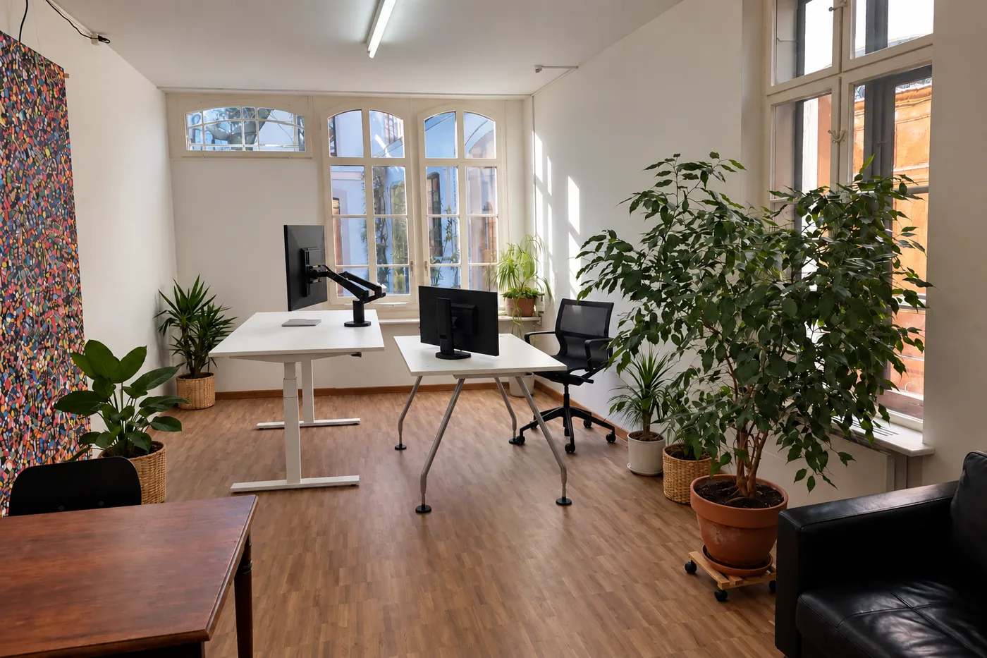 Büro von Konrad Sigl an der St. Alban-Vorstadt 53A in Basel: zwei Arbeitsplätze mit Monitoren vor hohen Sprossenfenstern, Zimmerpflanzen und Ledersofa